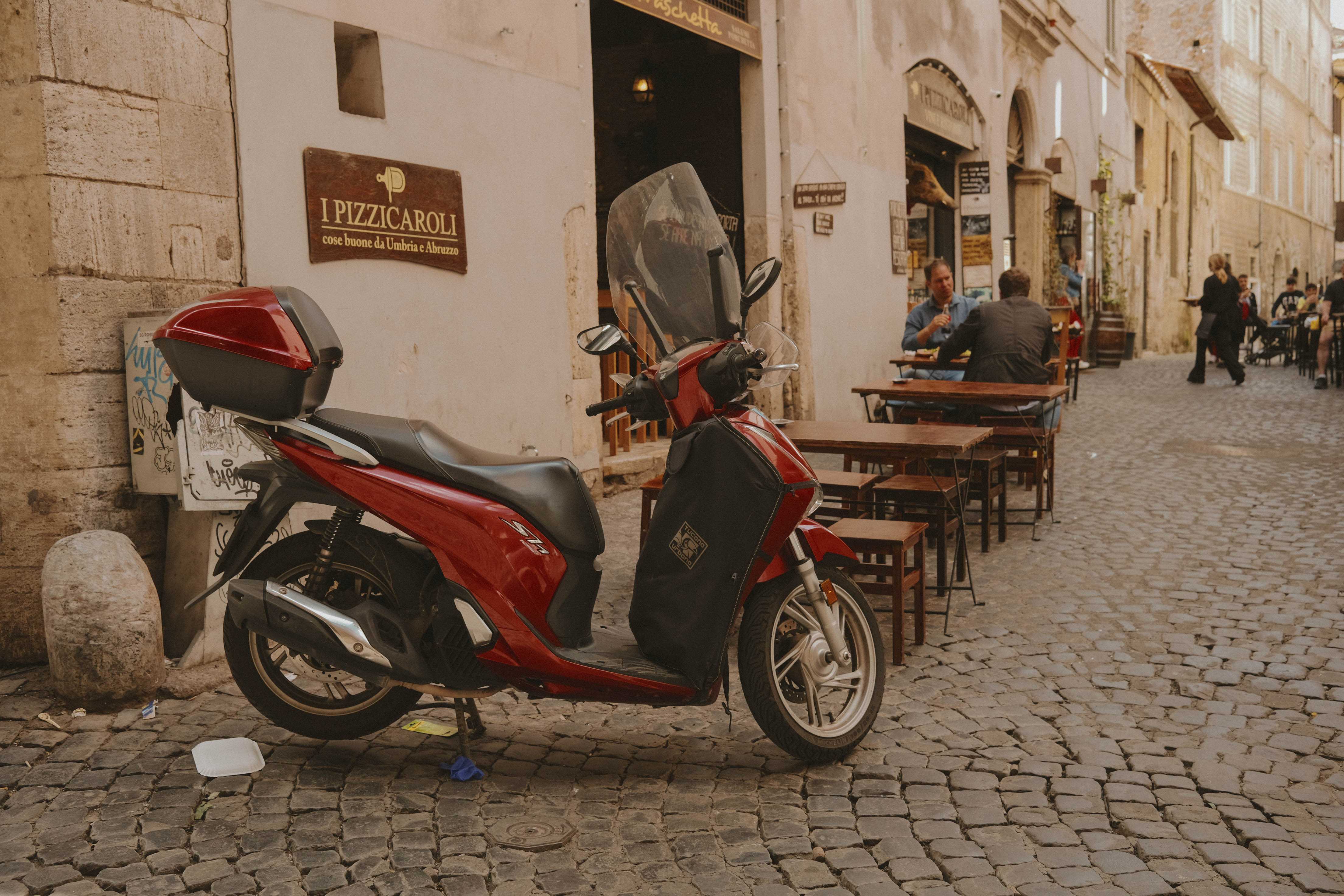A vespa-like scooter parked on the corner near a restaurant. It's on a cobbled street with several small tables.