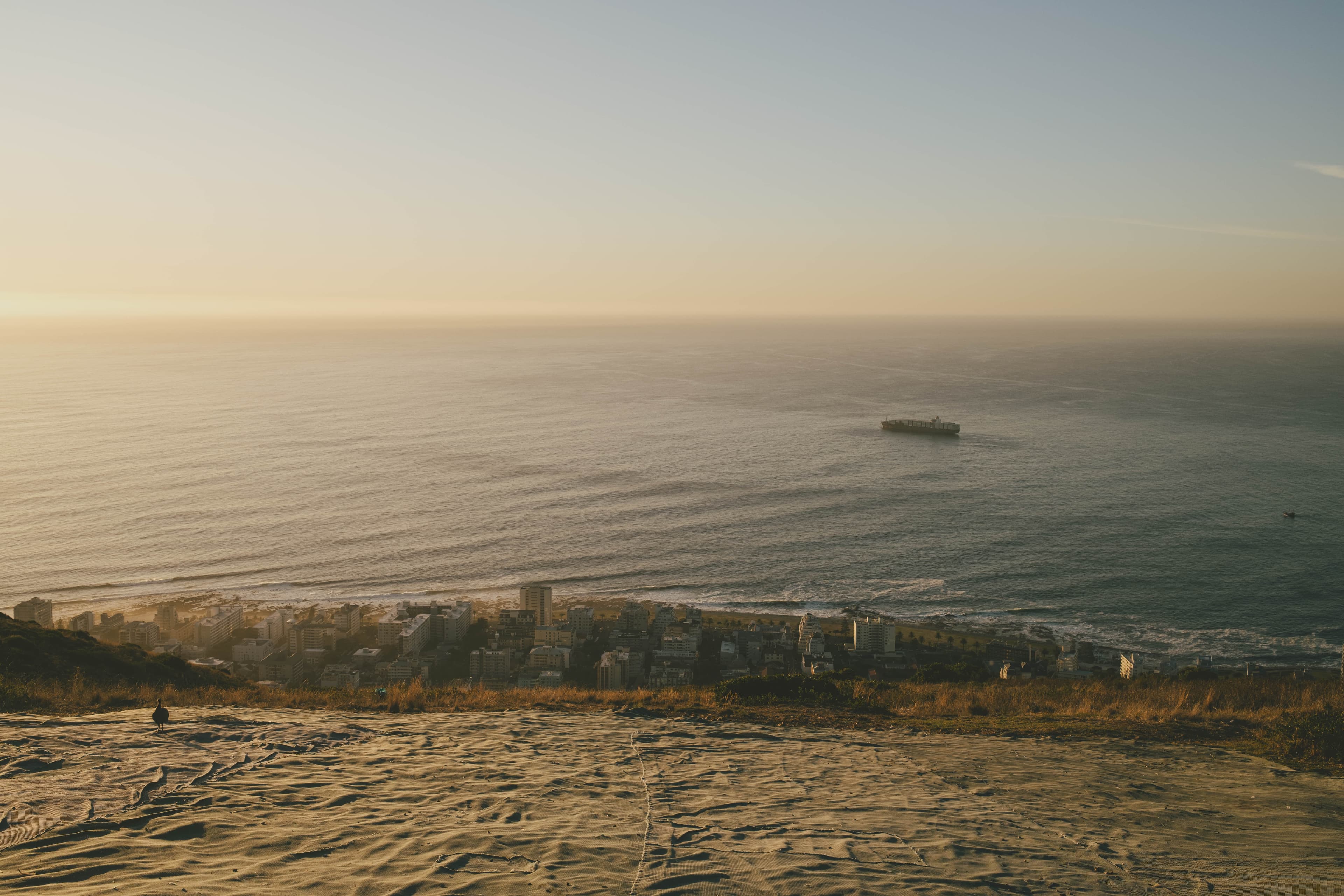 Looking down Signal Hill towards the ocean. The Green Point neighborhood is visible far below. In the sea, a container ship is passing by.