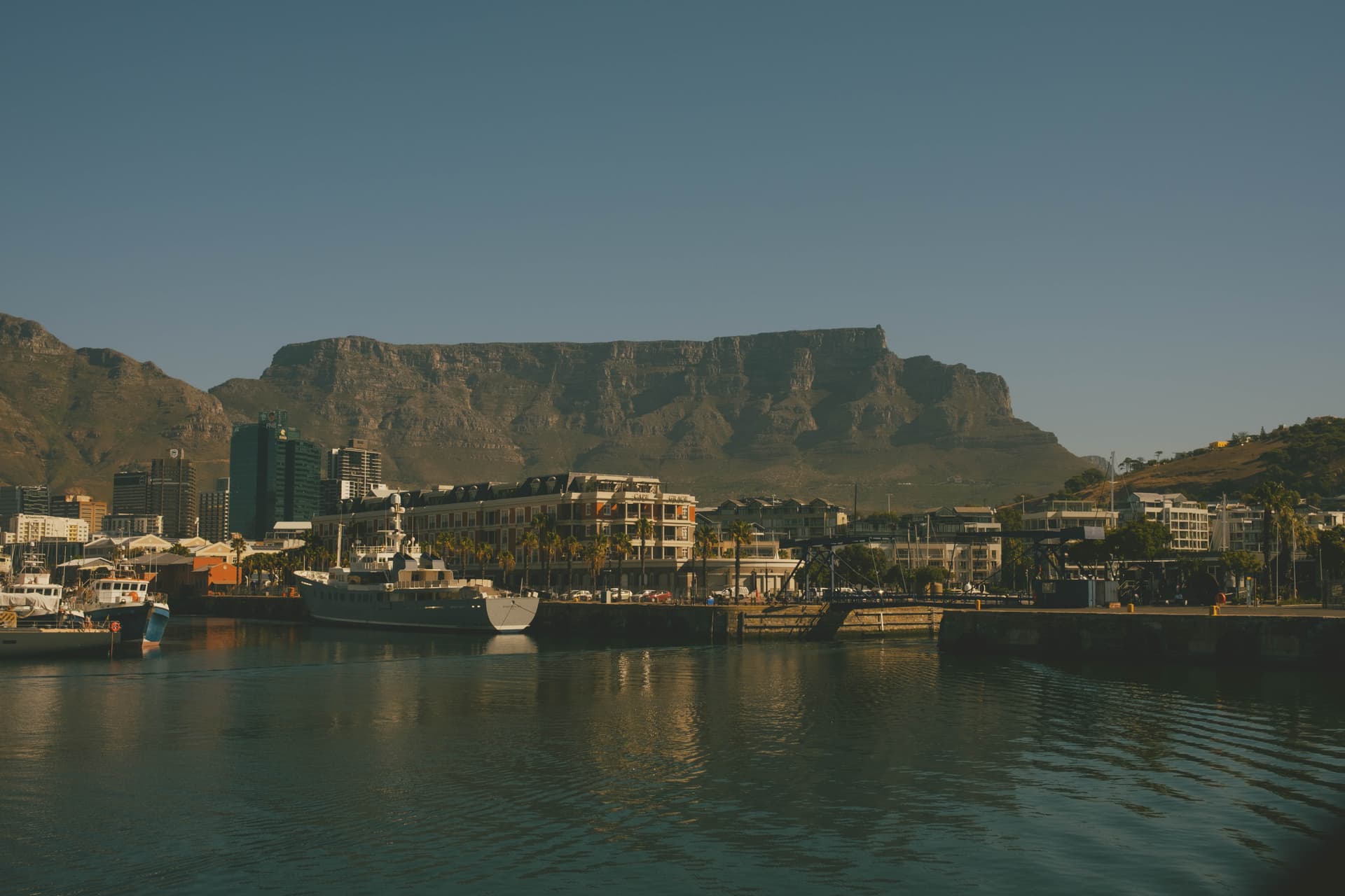 At the V&A Waterfront, looking across the harbor towards the city. There are several boats in the water. In the middle ground are buildings around the waterfront and the central business district. Table Mountain stands tall in the backdrop against a cloudless sky.