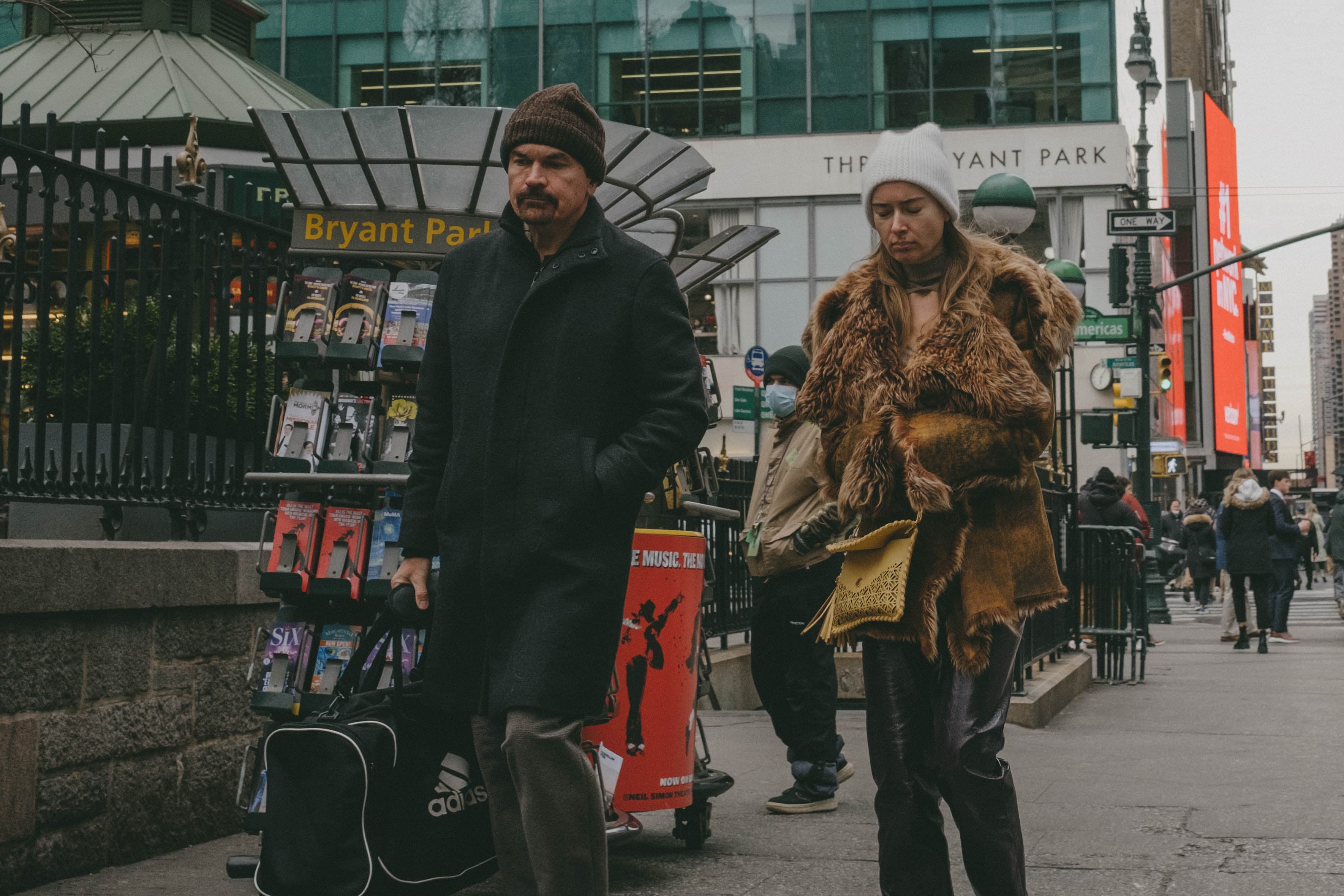 Two people walking, looking expressionless at the ground. One is a woman with a worn-down fur coat.