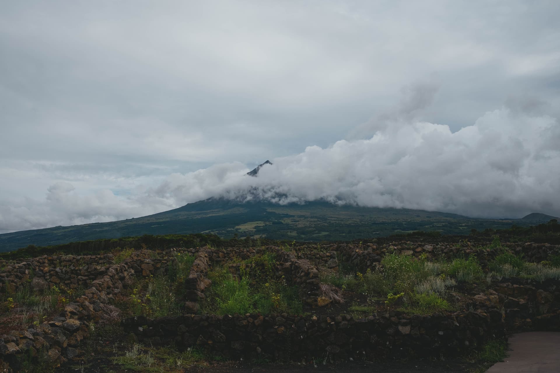 Pico volcano shrouded in dramatic clouds above a rocky lava field dotted with green vegetation under an overcast sky