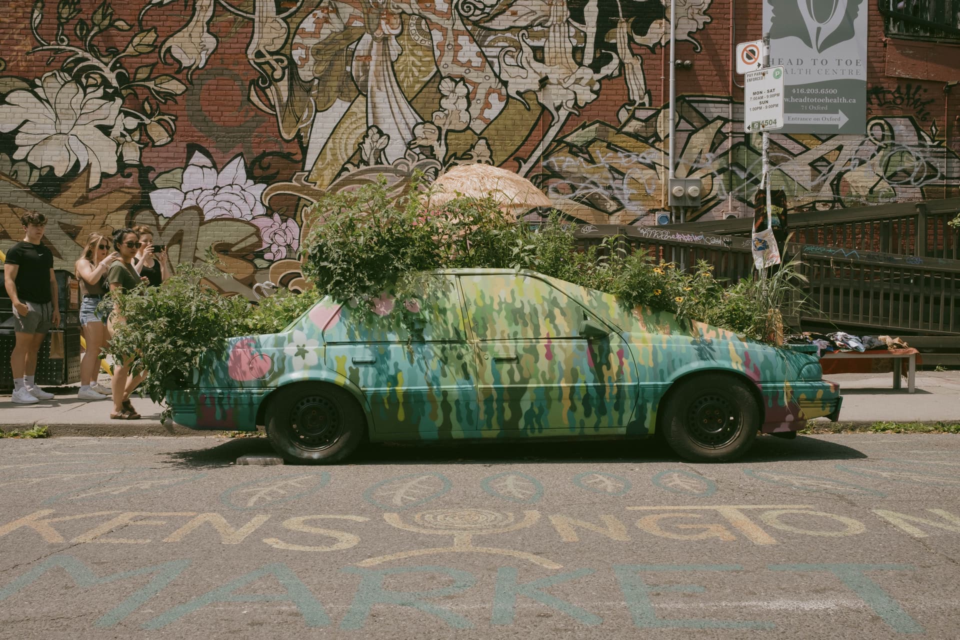 Heavily-painted car with plants growing out of every part of it. There are some onlookers behind it, and the word "Kensington" painted on the street.