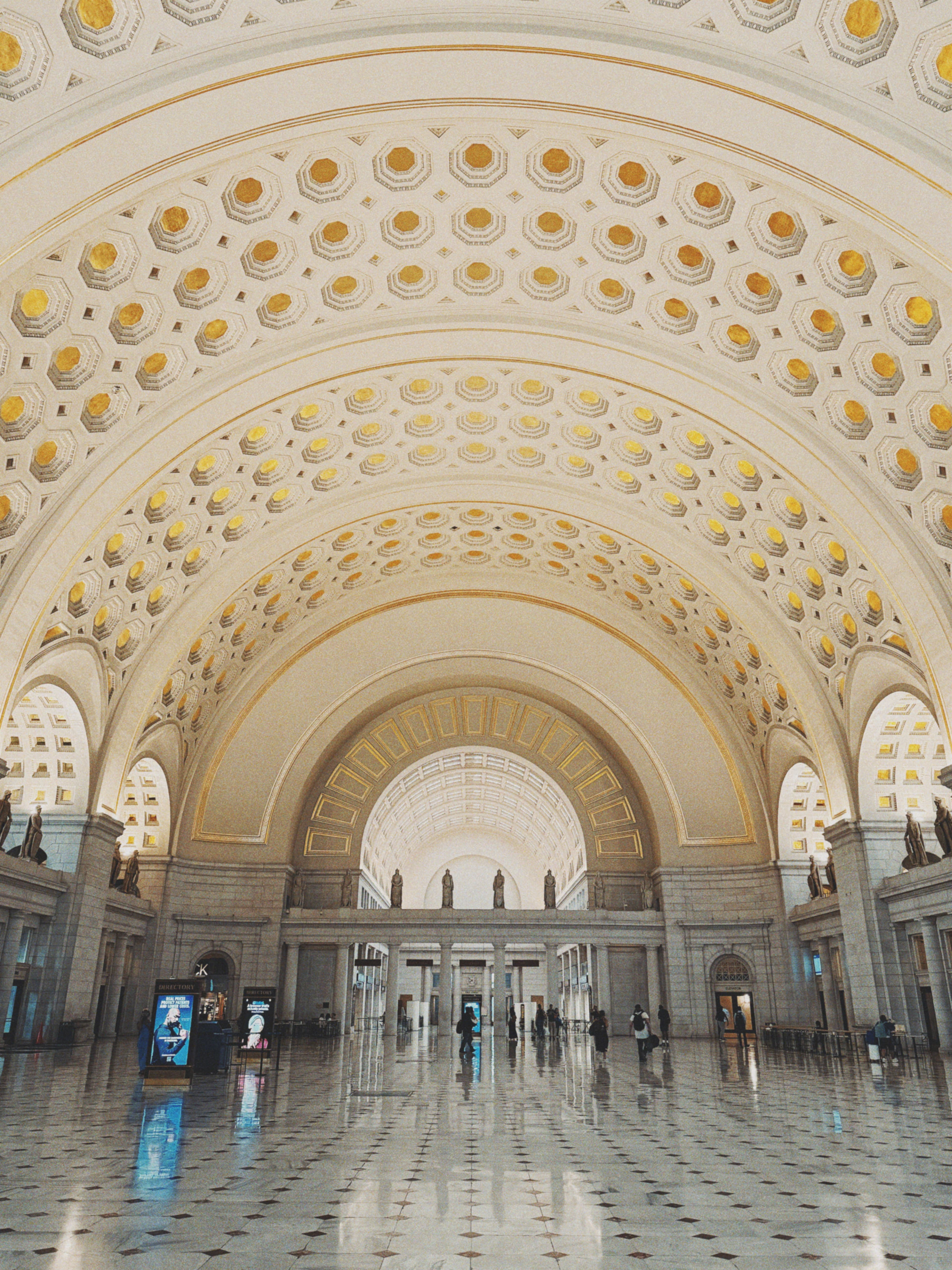 The grand interior hall of Union Station in Washington, D.C., with soaring barrel-vaulted ceilings decorated with golden coffers and ornate plasterwork, featuring classical architecture with arched openings and marble floors.