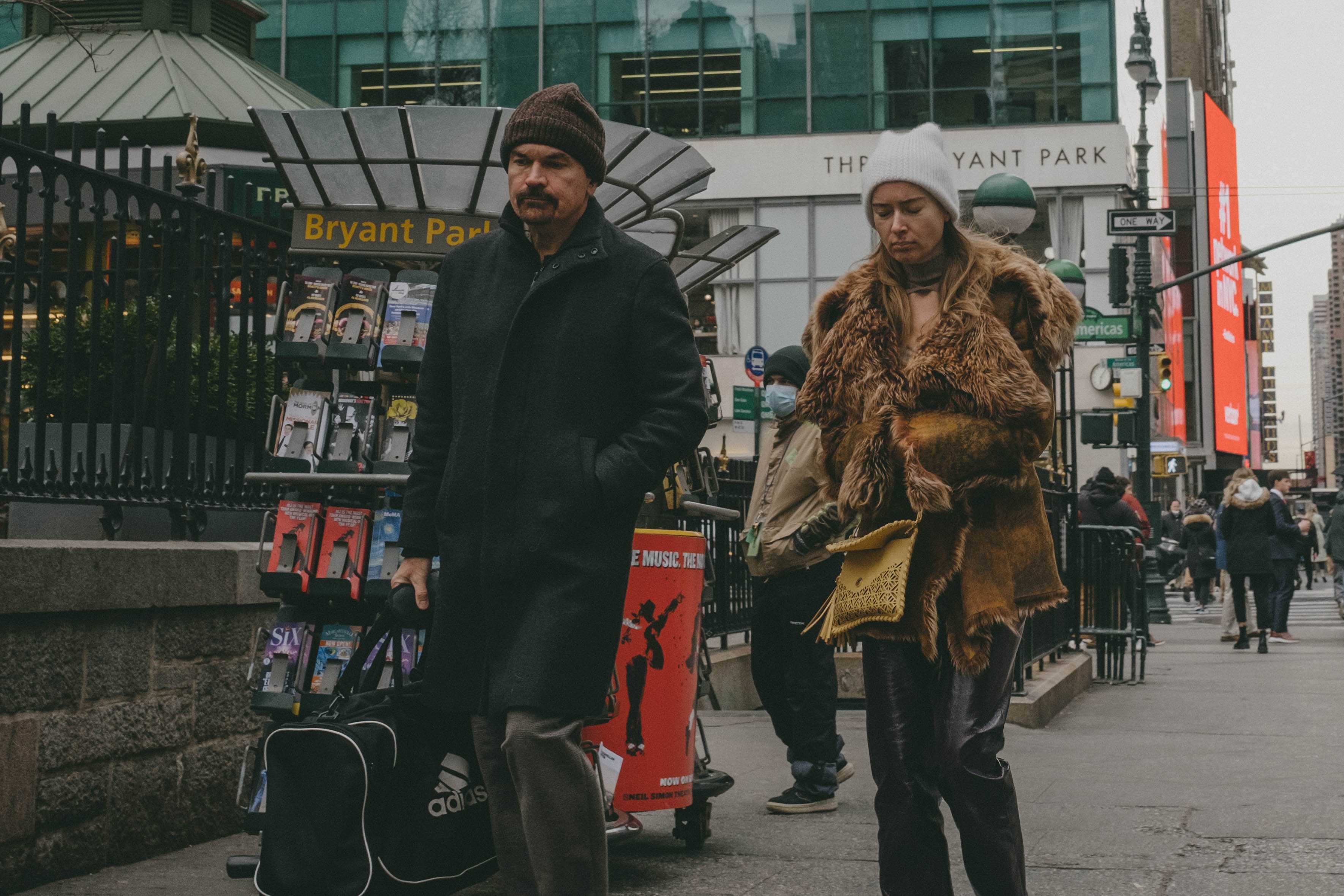Two people walking, looking expressionless at the ground. One is a woman with a worn-down fur coat.
