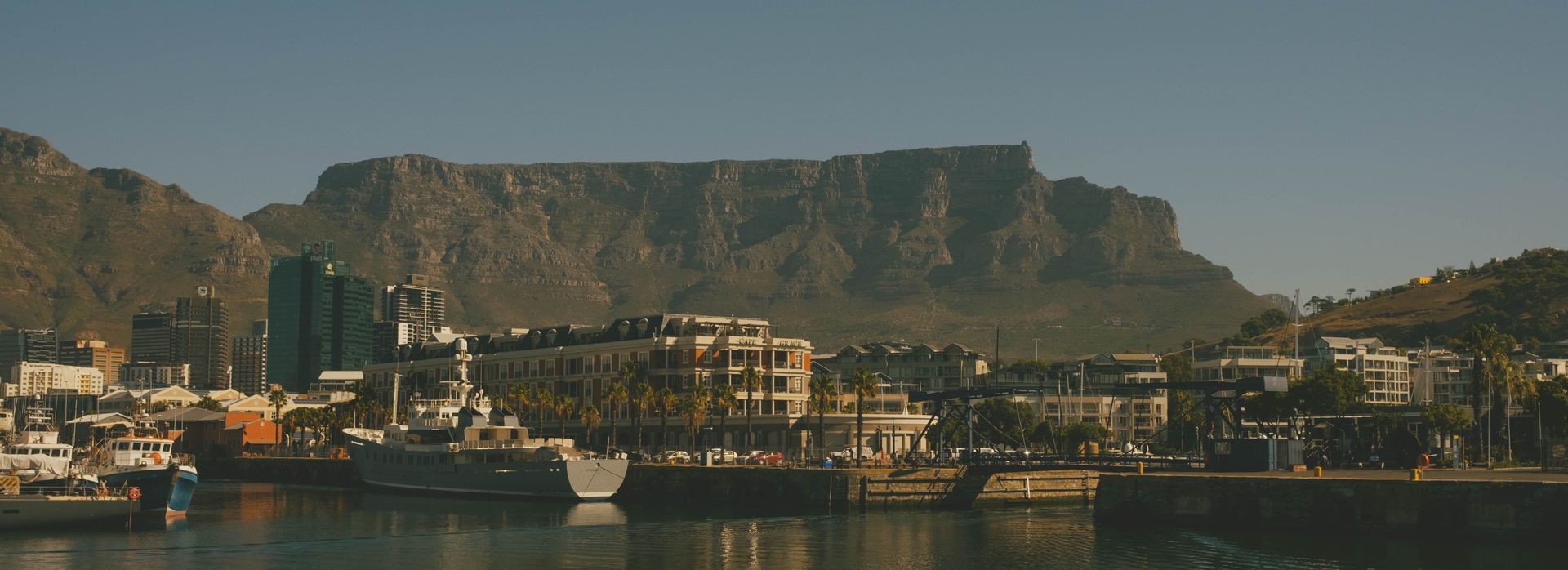 At the V&A Waterfront, looking across the harbor towards the city. There are several boats in the water. In the middle ground are buildings around the waterfront and the central business district. Table Mountain stands tall in the backdrop against a cloudless sky.