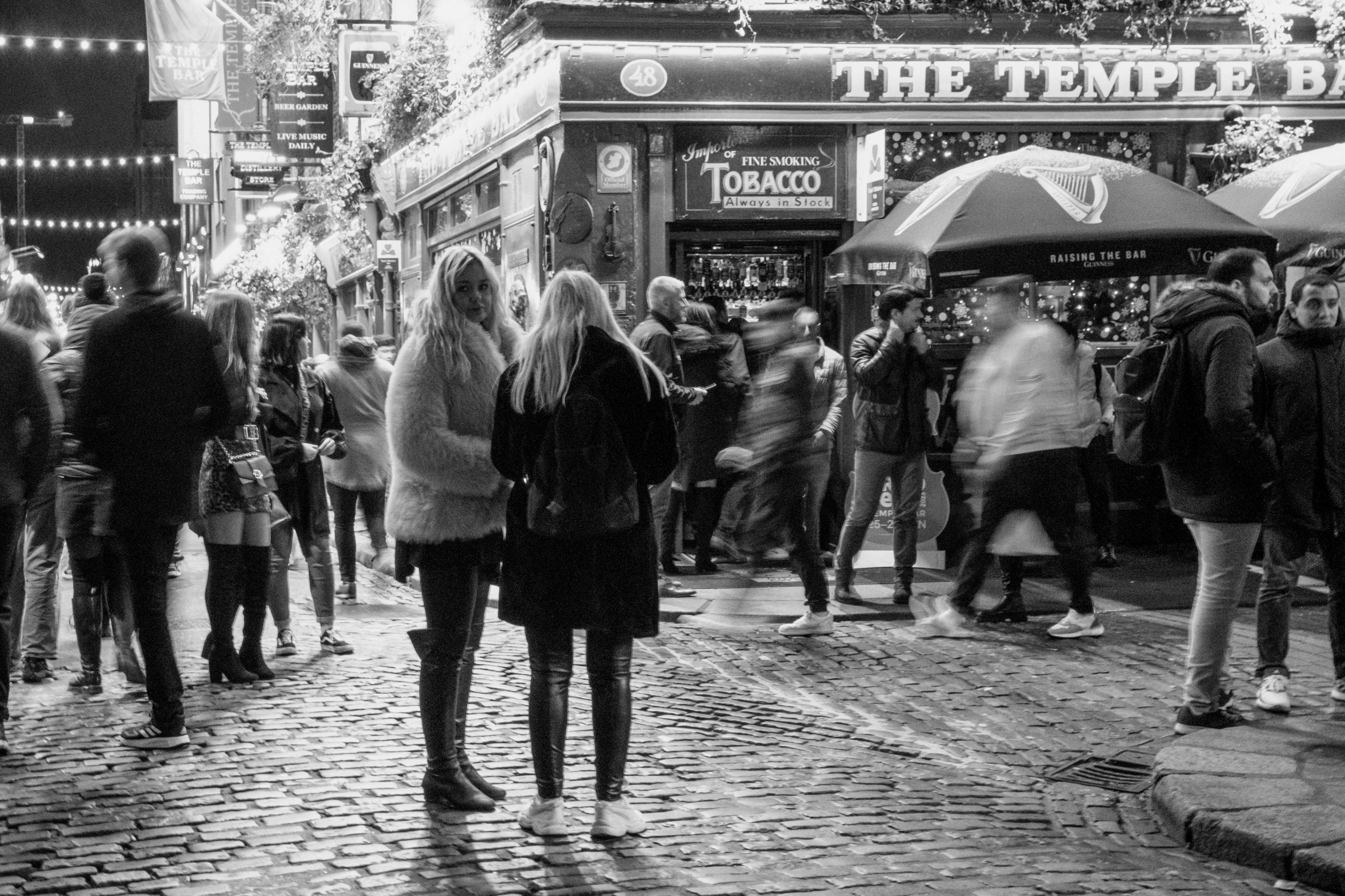 Two women in conversation outside of Temple Bar while the street is buzzing. One of them is looking at something behind her.