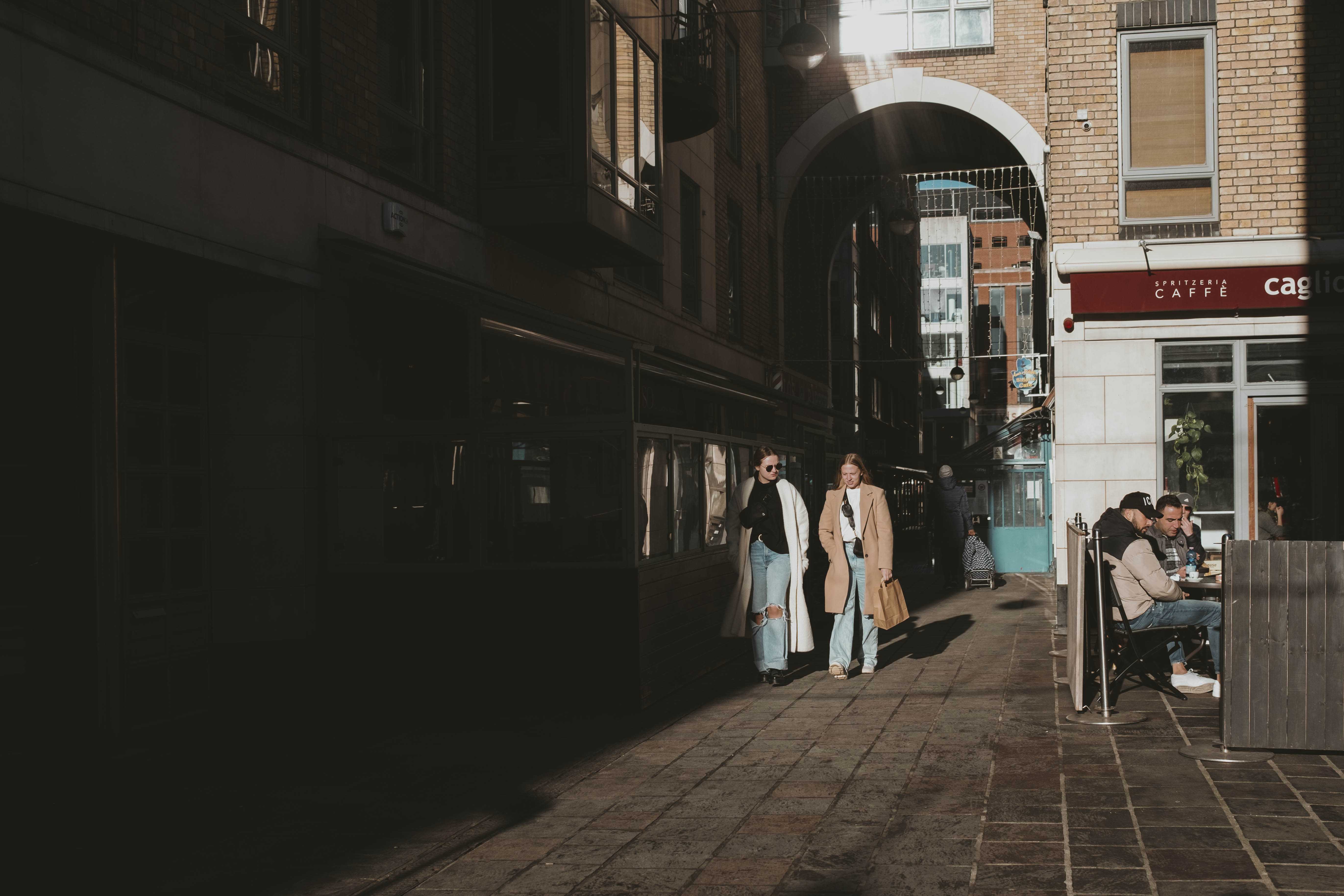 Two women walking through a corridor while harsh light illuminates them and the customers at the cafe on the right.