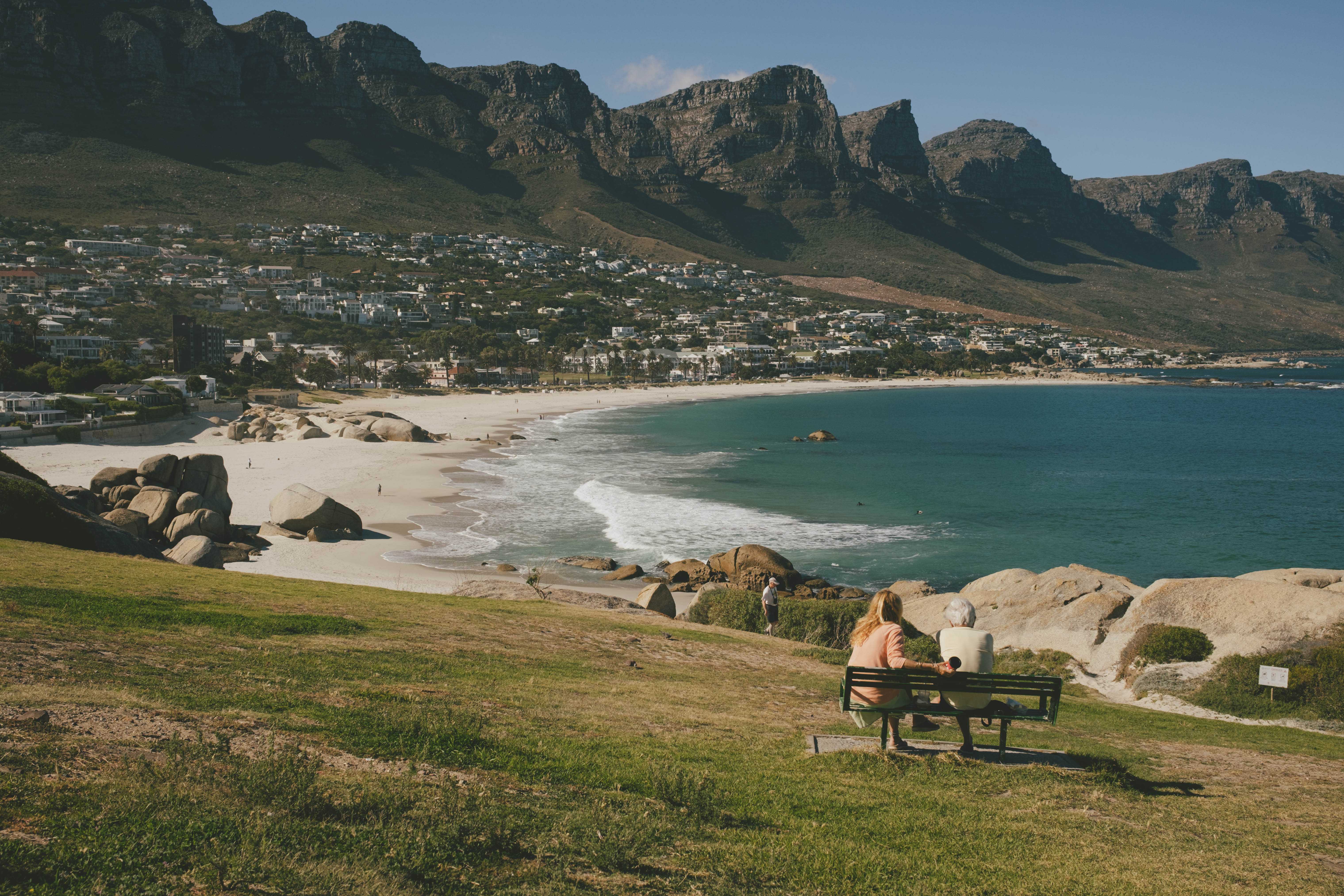 Two people sitting on a bench looking at the ocean in the Camps Bay neighborhood. The 12 Apostles mountains are in the background.