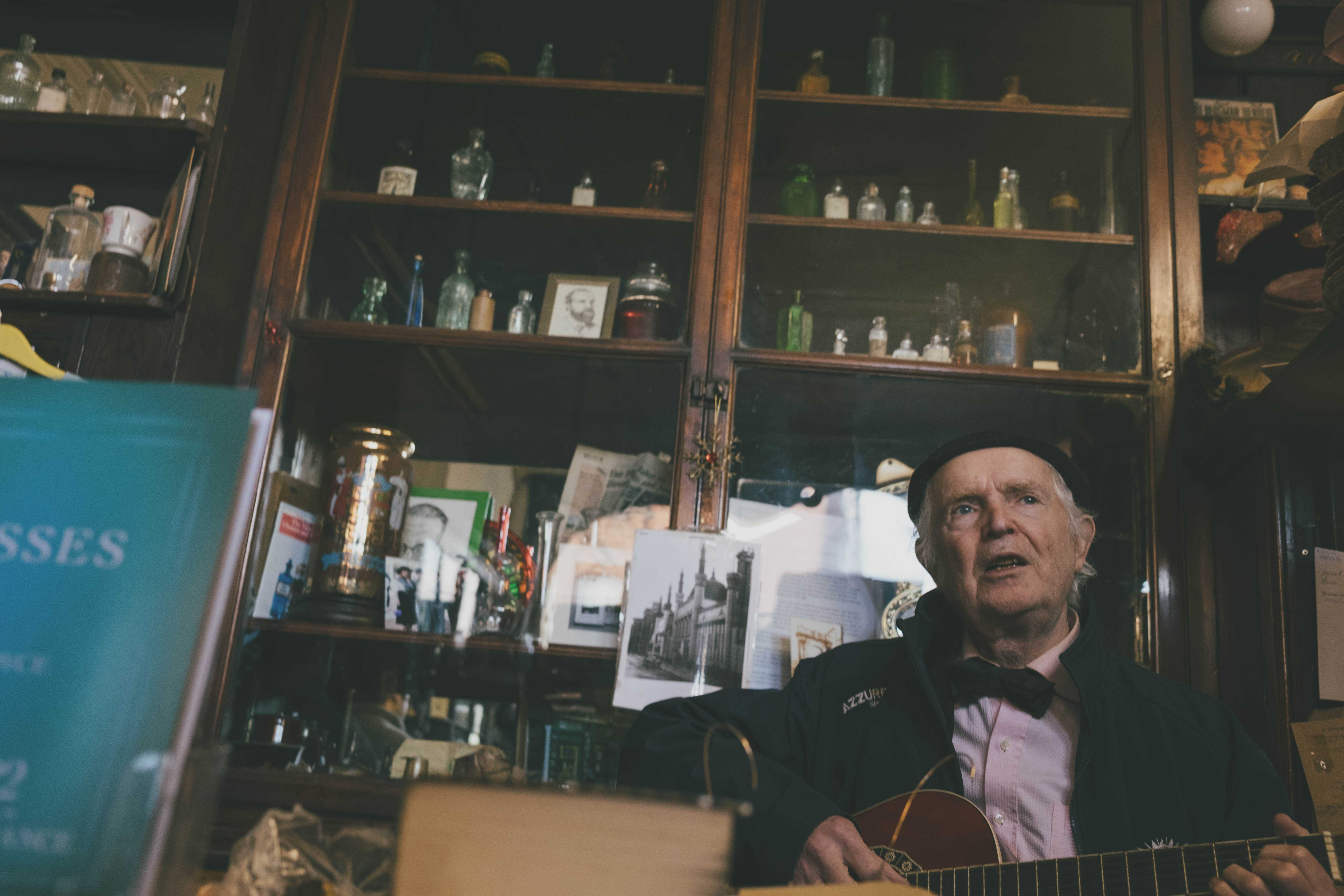 Sweny's proprietor playing his guitar and singing with old medicine bottles on a shelf behind him