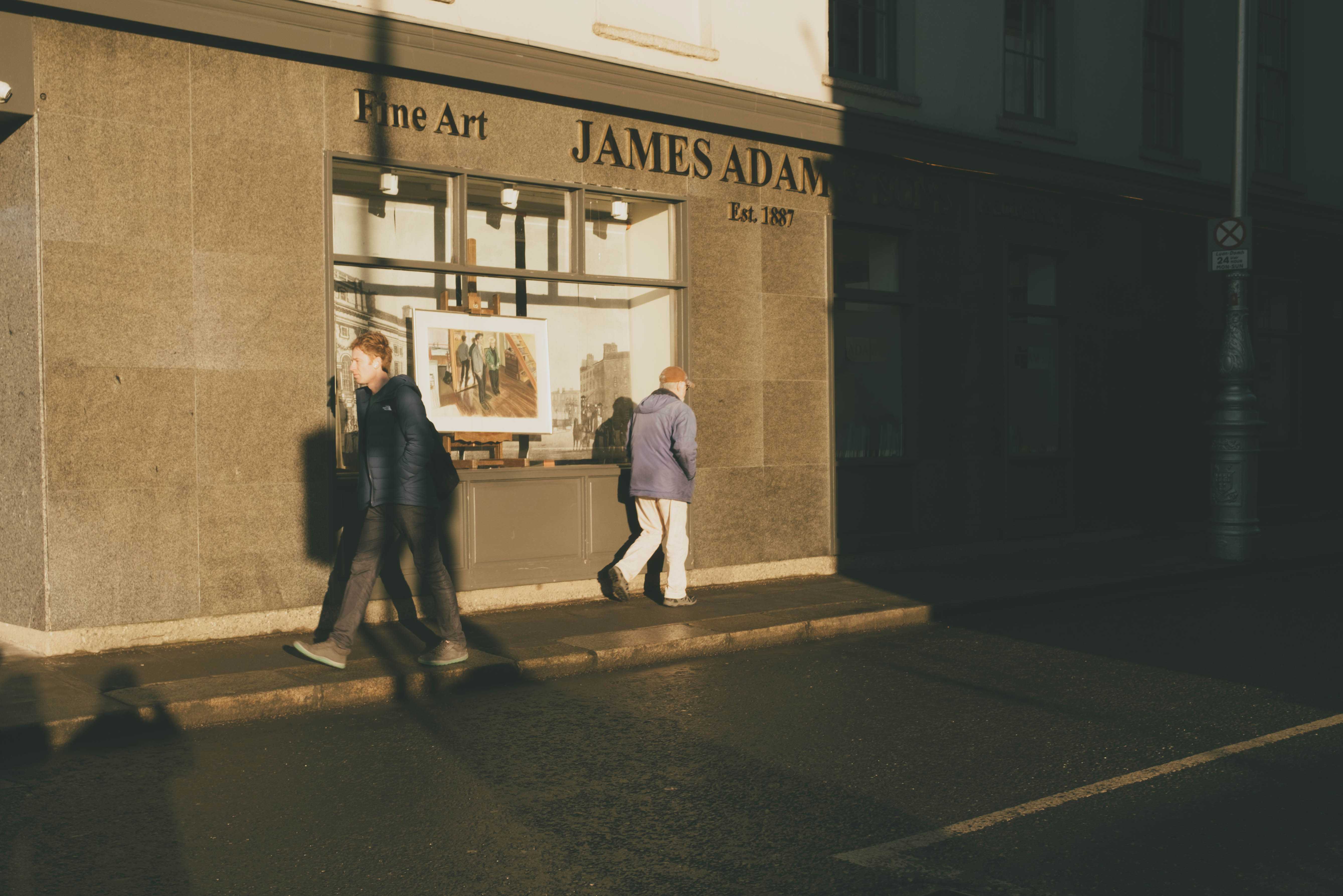 Two people walking in opposite directions, golden light against a wall with harsh shadows