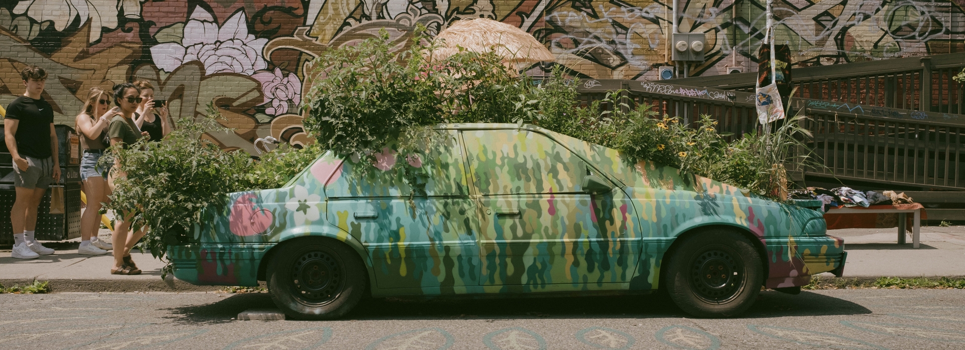 Heavily-painted car with plants growing out of every part of it. There are some onlookers behind it, and the word "Kensington" painted on the street.