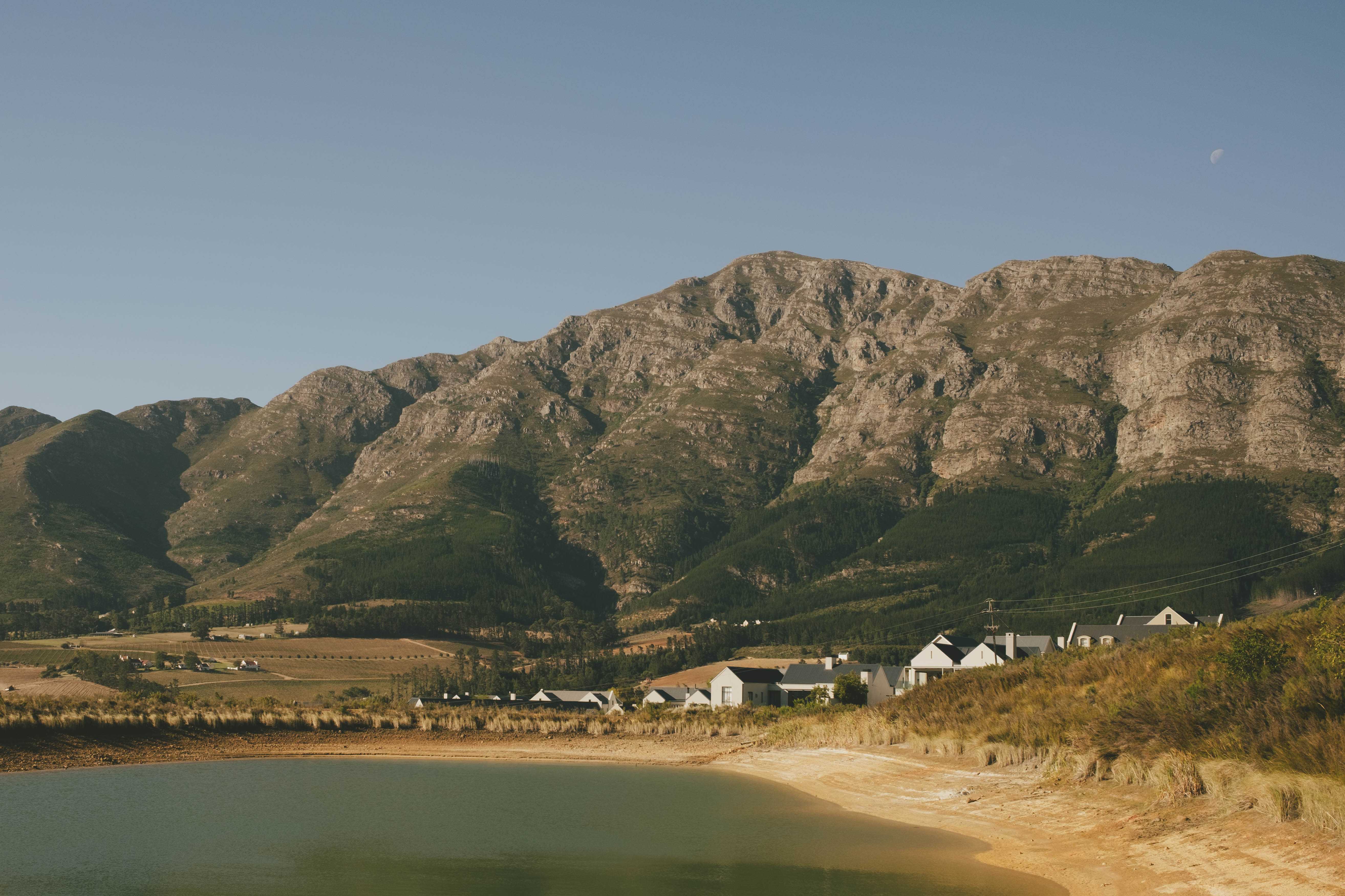 Homes against a mountain next a lake. The moon is visible in the late afternoon sun.