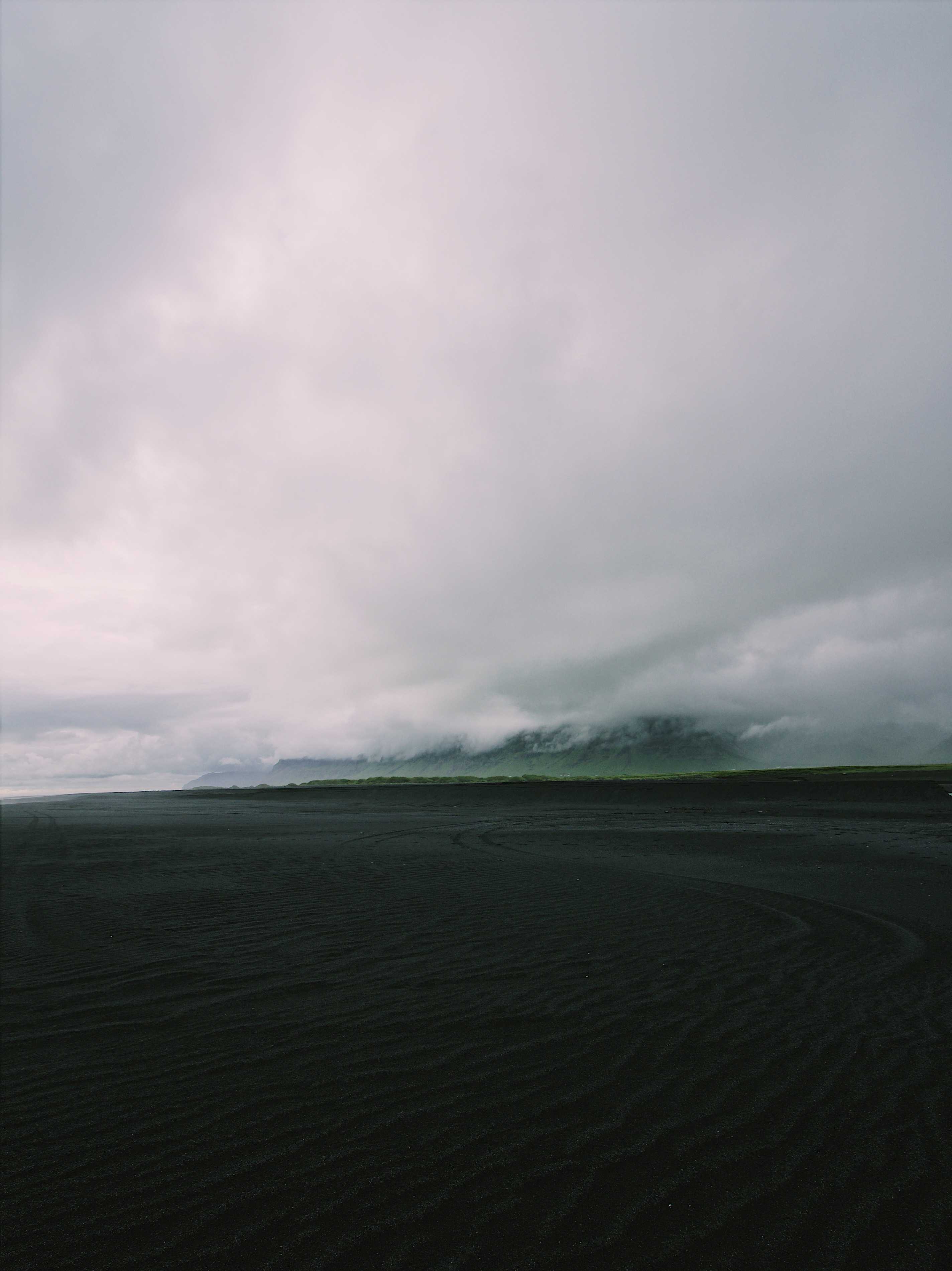 Black sand in the foreground with a mountain covered in clouds in the background
