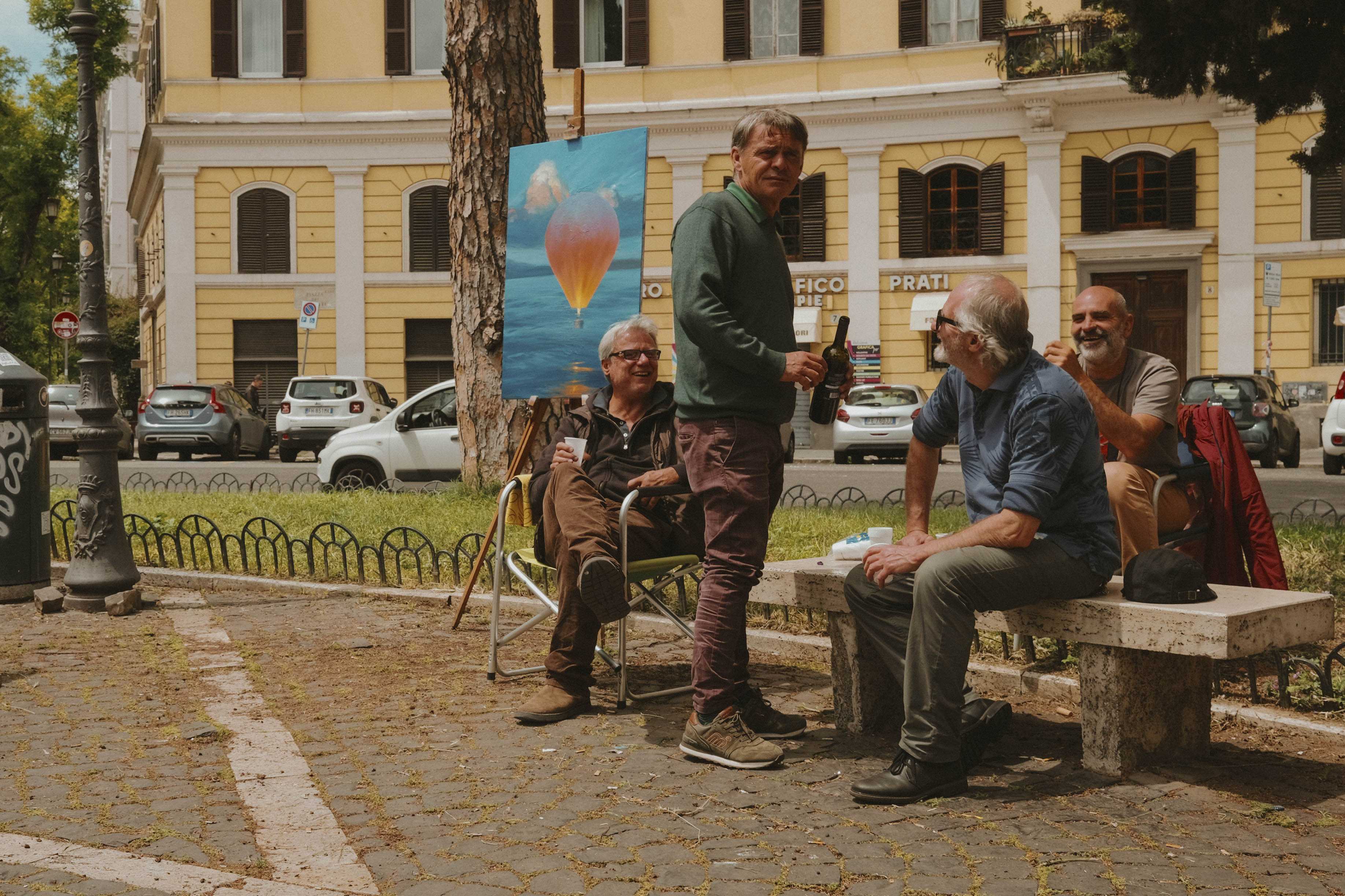 Group of older gentlemen laughing and sharing a bottle of wine at a park bench. One is turned around an seems to have noticed the camera