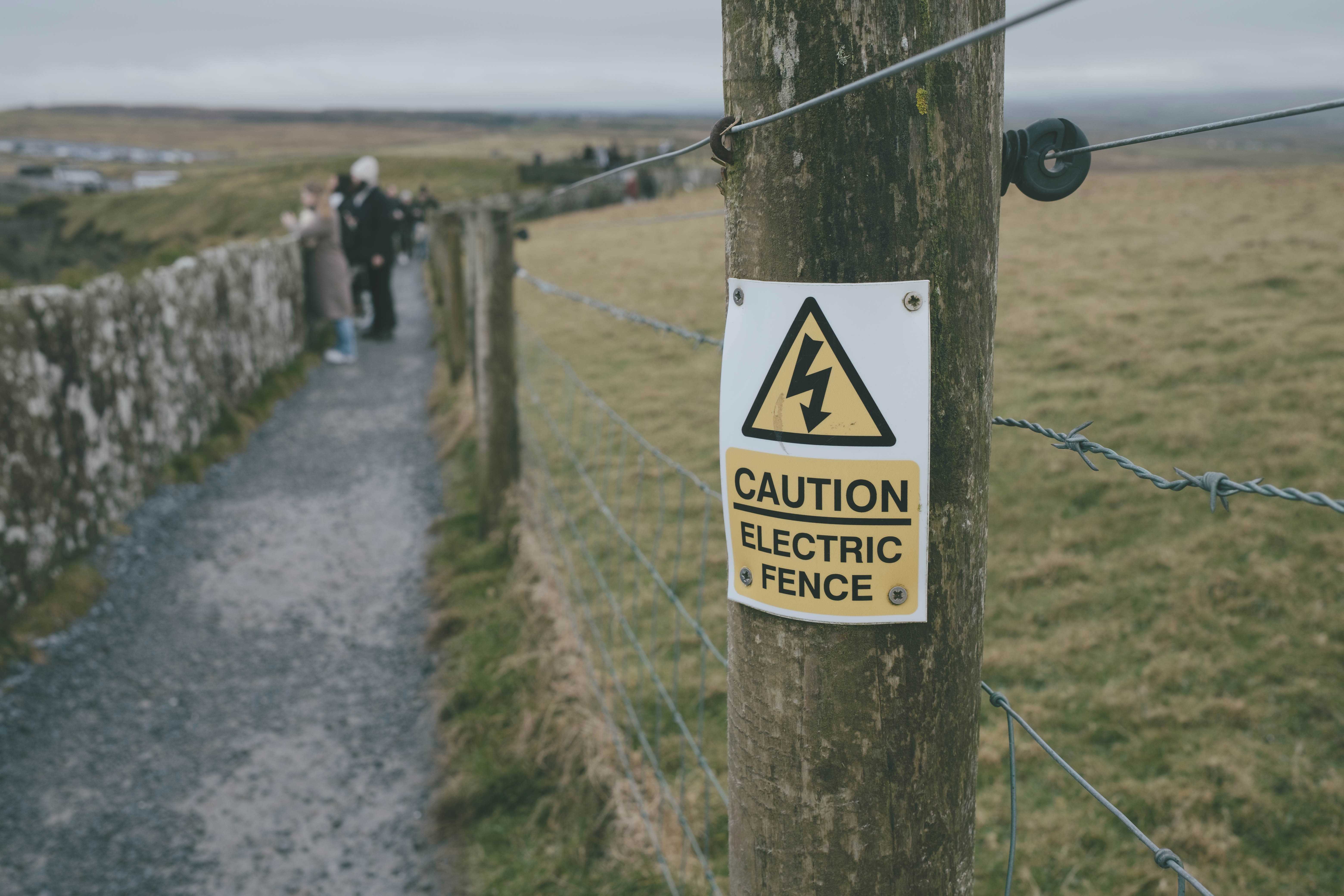 Narrow walkway between a fence and wall. The fence has a warning side that it is electrified