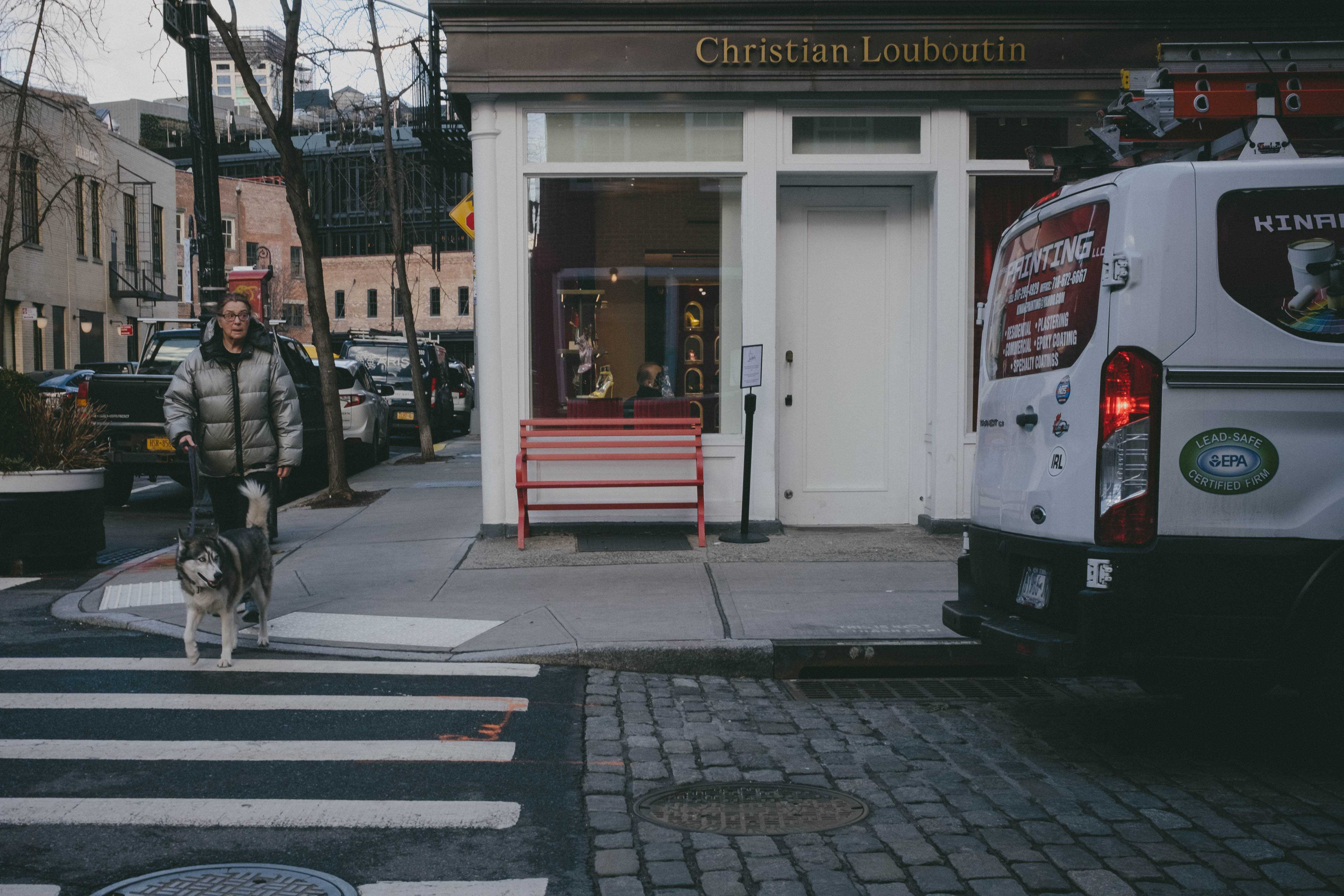 A woman crossing the street with her dog. Both of them are looking at a parked truck, as if it might start moving.